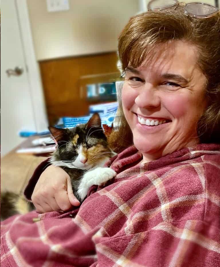 Woman smiling and cuddling her cat, showcasing the bond between humans and pets, emphasizing the importance of companionship for cats' well-being.
