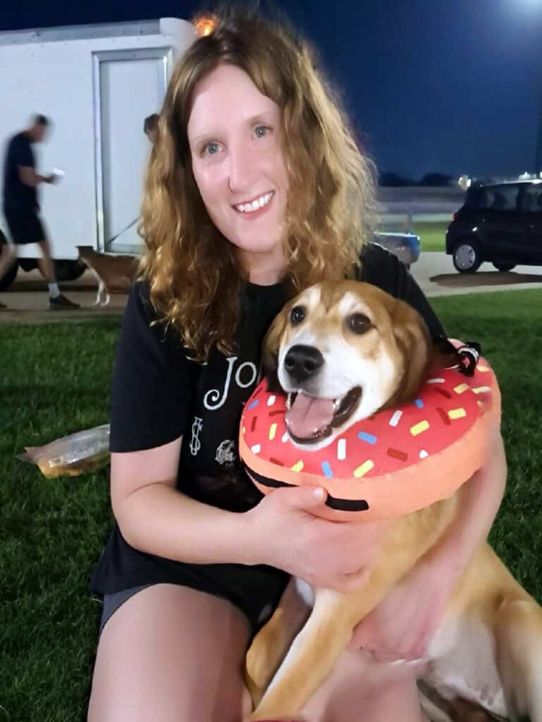 Young woman with curly red hair smiling and holding a happy foster dog with a donut-shaped collar at night, promoting dog fostering in Texas.